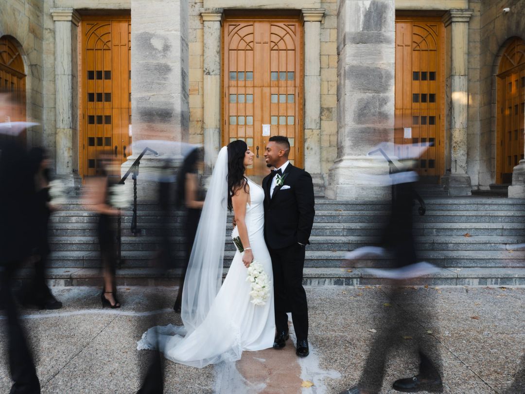 Bride and groom stand still on stone steps as the crowd blurs past